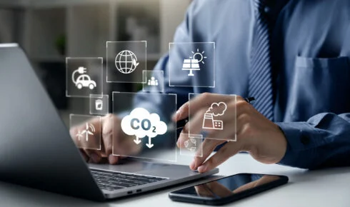 Scope 1 2 och 3: en fackperson arbetar på en bärbar dator, omgiven av digitala ikoner för CO2-utsläpp, en fabrik, förnybar energi och en elbil – en bild på den digitala spårning som ligger bakom företags utsläppsrapportering